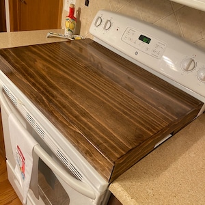 May include: A rectangular, dark stained wooden stove top cover. The cover is placed over a white GE stove. The wood grain is visible, and the cover has a glossy finish. A white towel with a red and blue design hangs on the stove.