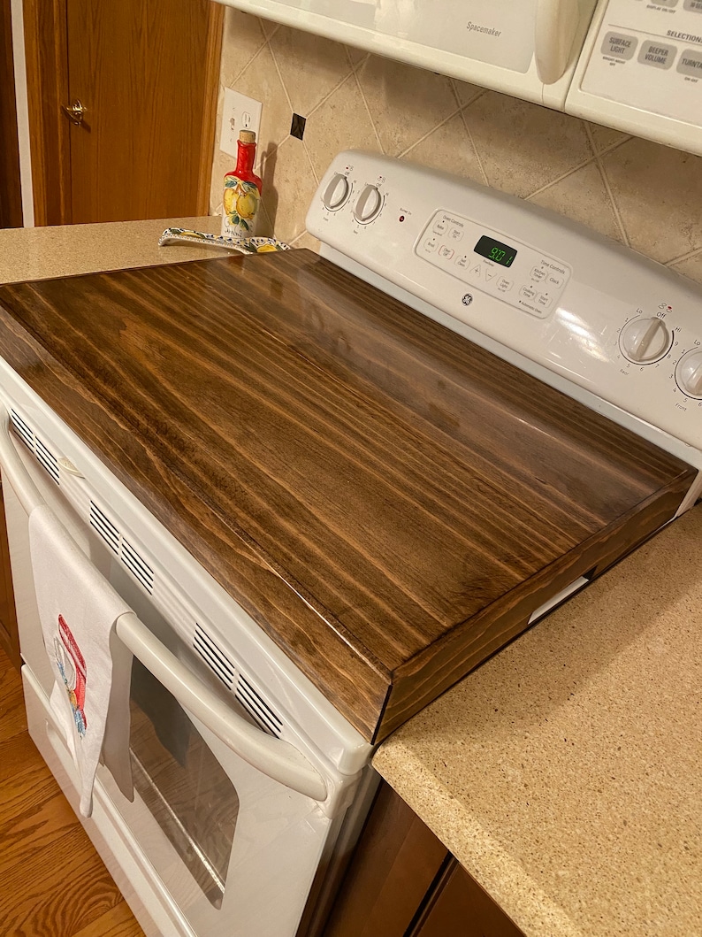 May include: A rectangular, dark stained wooden stove top cover. The cover is placed over a white GE stove. The wood grain is visible, and the cover has a glossy finish. A white towel with a red and blue design hangs on the stove.
