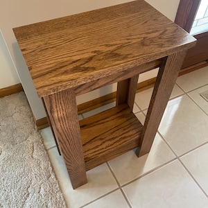 May include: A rectangular wooden side table with a medium brown finish and visible wood grain. The table features a flat top surface, four straight legs, and a lower shelf positioned between the legs, providing an open storage area.