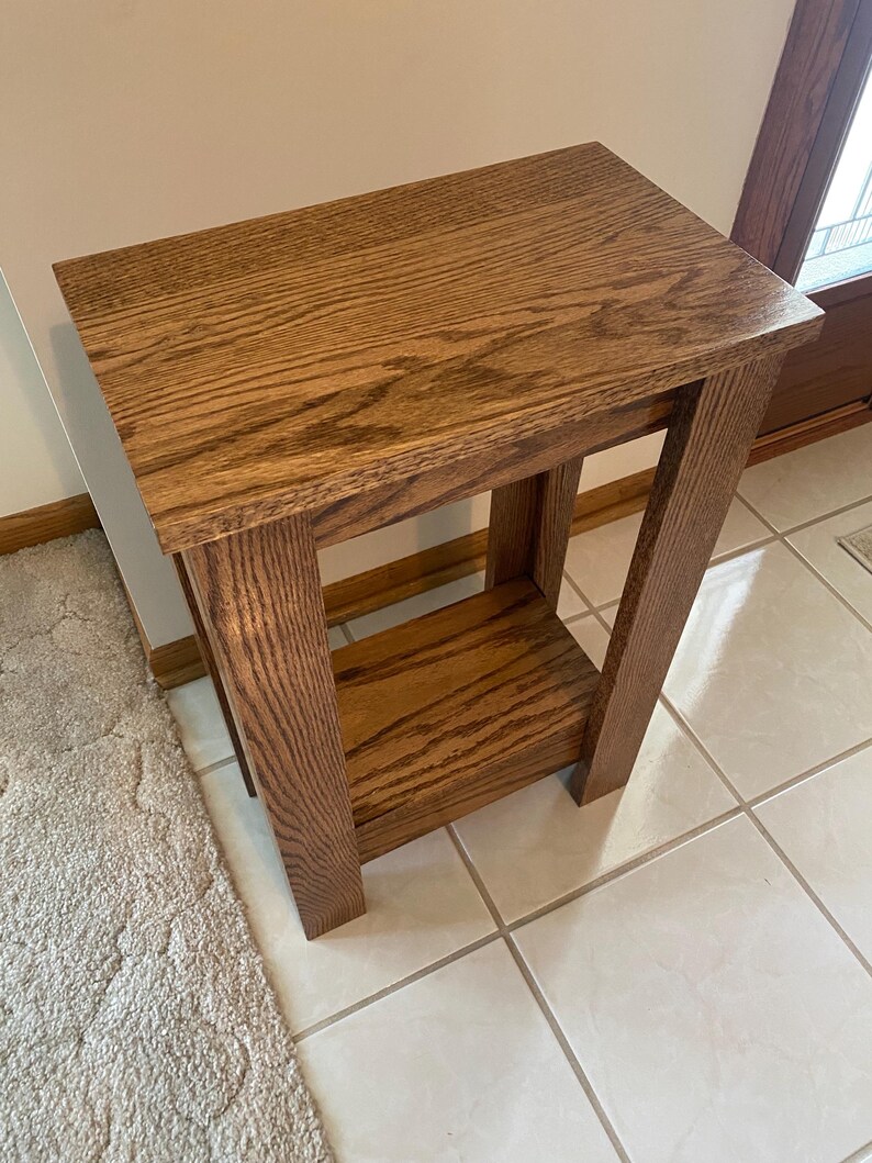 May include: A rectangular wooden side table with a medium brown finish and visible wood grain. The table features a flat top surface, four straight legs, and a lower shelf positioned between the legs, providing an open storage area.