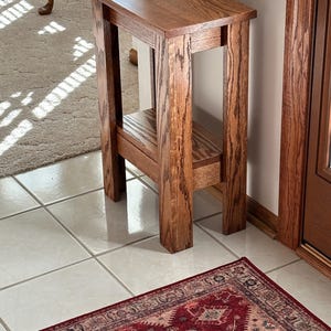 May include: A tall, narrow rectangular side table constructed from medium-brown stained wood with a visible grain pattern. The table features four straight legs, a flat rectangular top surface, and a lower shelf positioned between the legs.
