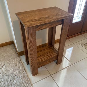 May include: A rectangular wooden side table featuring a medium brown wood grain finish. The table has a flat top surface, four sturdy vertical legs, and a lower shelf positioned between the legs, providing an open structure with clean lines.