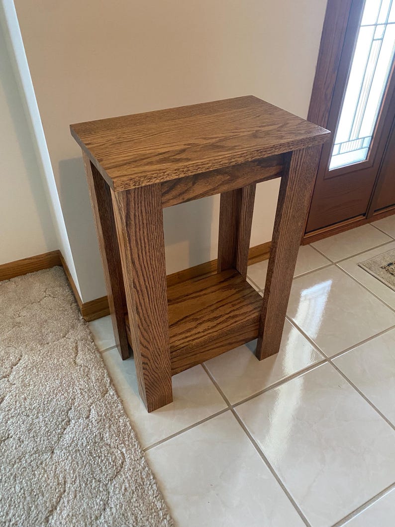 May include: A rectangular wooden side table featuring a medium brown wood grain finish. The table has a flat top surface, four sturdy vertical legs, and a lower shelf positioned between the legs, providing an open structure with clean lines.