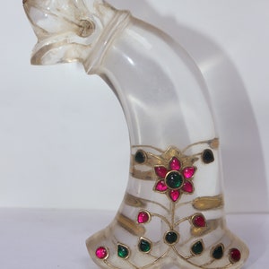 May include: A clear, curved decorative object with a dog-like head. The base features a floral design with red and green gemstones, accented with gold-coloured metal. The object has a pearlescent appearance.