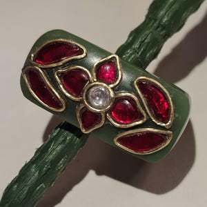 Puede incluir: Un anillo verde con un diseño floral. El anillo presenta una flor con una piedra central transparente y pétalos rojos, contorneados en oro. Acentos rojos en forma de hoja también están contorneados en oro.