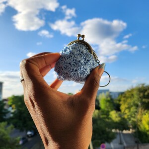Könnte beinhalten: Eine hellblaue gehäkelte Geldbörse mit einem Messingverschluss und einem Metallring, der an einer Kette befestigt ist. Die Börse wird in einer Hand vor einem blauen Himmel gehalten.