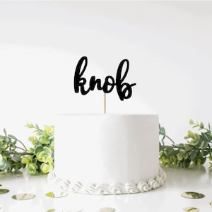 May include: A white cake with a black cursive "knob" topper. The cake is decorated with white frosting and a scalloped edge. The cake is sitting on a white surface with gold confetti.