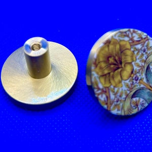 May include: Two cabinet knobs, one with a brass finish and the other with a floral pattern. The floral knob has a yellow flower with green and white leaves on a white background.