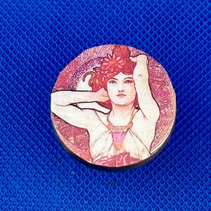 May include: Round wooden coaster featuring an Art Nouveau style illustration of a woman with red hair and a floral headdress. The artwork is set against a pink and cream background with decorative patterns. The coaster has a dark brown edge.
