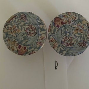 May include: A pair of round, floral patterned earrings. The earrings are made of a white fabric with a blue, red, and yellow floral design.