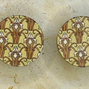 May include: A pair of round, gold-toned earrings with a floral design. The design features white flowers with brown stems and leaves on a yellow background.