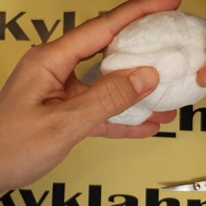 May include: A small, round, white plush fabric sphere being held and manipulated. The object has a soft, fuzzy texture and is constructed from multiple stitched panels. A spool of white thread and a pair of metal scissors are visible nearby.