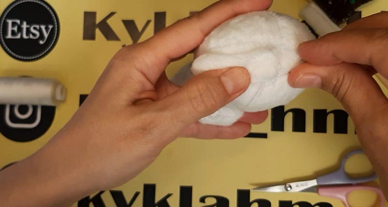 May include: A small, round, white plush fabric sphere being held and manipulated. The object has a soft, fuzzy texture and is constructed from multiple stitched panels. A spool of white thread and a pair of metal scissors are visible nearby.