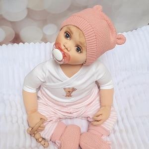 Puede incluir: Una muñeca bebé con un gorro de punto rosa con detalles de orejas, un body blanco con un gráfico de oso de peluche y pantalones a rayas rosas. La muñeca tiene un chupete y botines de punto rosa. Una sonaja de madera está en su mano.