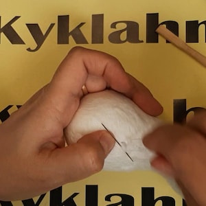 May include: A white, rounded, soft-textured object being worked on with a thin metal needle. A long, slender wooden dowel rests nearby on a yellow surface featuring black text and logos, including the Etsy and Instagram icons.
