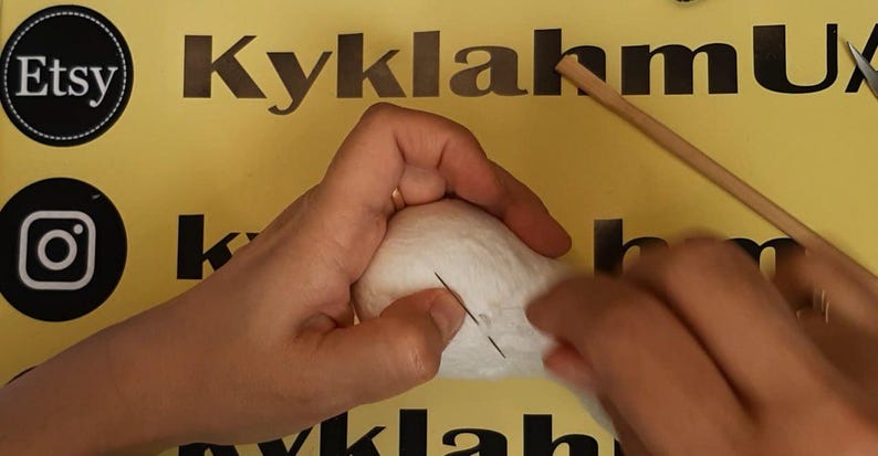 May include: A white, rounded, soft-textured object being worked on with a thin metal needle. A long, slender wooden dowel rests nearby on a yellow surface featuring black text and logos, including the Etsy and Instagram icons.