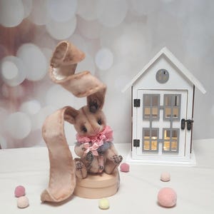 May include: A beige fabric rabbit figurine with one long, curled ear and one upright ear, wearing a pink ruffled collar and a small silver charm. The rabbit sits on a round wooden base next to a white miniature house with gray window frames.