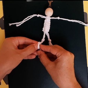 May include: A small figurine crafted from white cord with a round wooden bead for a head. The figure has outstretched arms and legs formed by knotted cord, with a metal clip attached to the top of the head. The design is simple and linear.