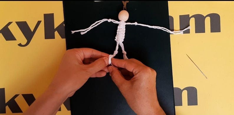 May include: A small figurine crafted from white cord with a round wooden bead for a head. The figure has outstretched arms and legs formed by knotted cord, with a metal clip attached to the top of the head. The design is simple and linear.