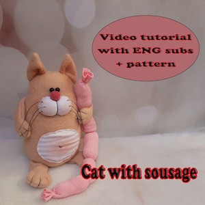May include: A plush cat toy in shades of peach and white, holding a pink sausage-shaped toy. The cat has black whiskers, a pink nose, and a white belly. Text on the image reads "Video tutorial with ENG subs + pattern" and "Cat with sousage."