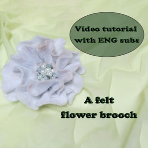 Op de afbeelding: Een lichtblauwe viltbloem broche met golvende bloemblaadjes en een cluster van witte en blauwe kralen in het midden. De afbeelding bevat de tekst: Video tutorial with ENG subs en A felt flower brooch.