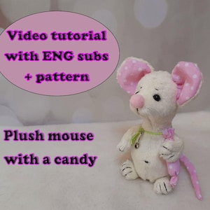 May include: A plush white mouse with pink polka-dot ears and a pink nose. The mouse holds a pink candy and wears a green collar. Text on the image reads "Video tutorial with ENG subs + pattern" and "Plush mouse with a candy."