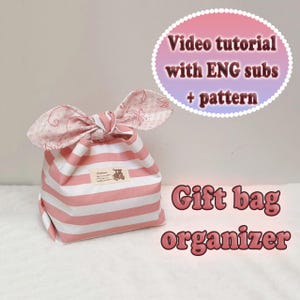 May include: A pink and white striped gift bag organizer with a bow-shaped top. The bag has a small label and the text "Video tutorial with ENG subs + pattern" and "Gift bag organizer".