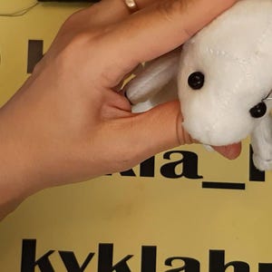 May include: A small, white plush toy with black bead eyes being stitched with a needle and thread. The fabric has visible seams across the head and body, creating a soft, stuffed animal form. The toy is held firmly while the needle is inserted.