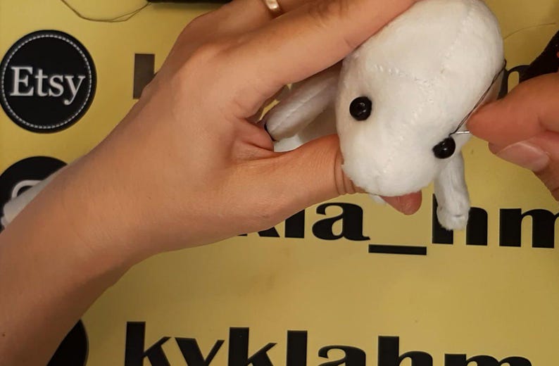 May include: A small, white plush toy with black bead eyes being stitched with a needle and thread. The fabric has visible seams across the head and body, creating a soft, stuffed animal form. The toy is held firmly while the needle is inserted.