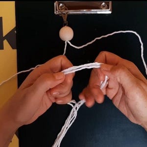 May include: A white wooden bead attached to a metal clip on a black clipboard, with white cotton cord being manipulated by hands to create a macrame knot. The cord is thin and white, forming multiple strands that are being looped and pulled together.