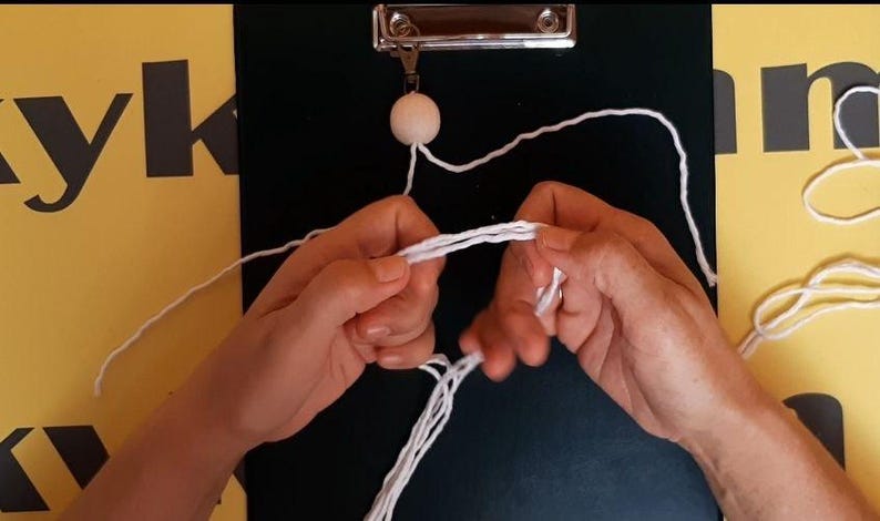 May include: A white wooden bead attached to a metal clip on a black clipboard, with white cotton cord being manipulated by hands to create a macrame knot. The cord is thin and white, forming multiple strands that are being looped and pulled together.