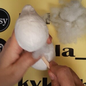 May include: A white plush fabric shape being stuffed with white polyester fiberfill using a wooden dowel. The object is held against a yellow background featuring black Etsy and Instagram logos and partial black text.