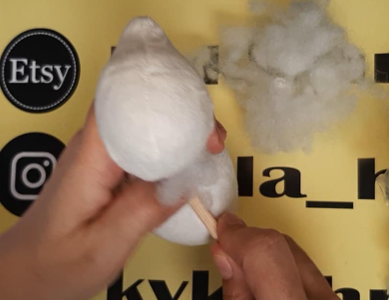 May include: A white plush fabric shape being stuffed with white polyester fiberfill using a wooden dowel. The object is held against a yellow background featuring black Etsy and Instagram logos and partial black text.