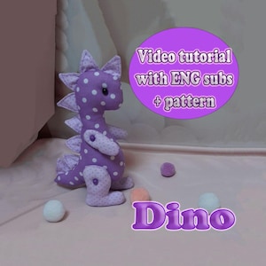 May include: A handcrafted purple dinosaur soft toy with white spots, featuring a spiky back and small arms. The image includes the text "Video tutorial with ENG subs + pattern" and the word "Dino". Several small, colourful pom-poms are scattered around the toy.