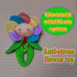 May include: A handmade flower-shaped plush toy with a smiling face and colorful petals. The petals are various colors and patterns. The toy has green leaves and a green body. Text on the image says "Video tutorial with ENG subs + pattern" and "Anti-stress flower toy."