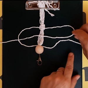 May include: A white macrame cord is being knotted around a central vertical strand, which is secured to a black clipboard. A single round, light-colored wooden bead is threaded onto the cord, positioned above a silver-toned metal lobster clasp.