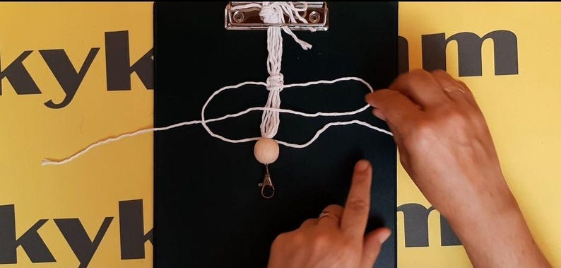 May include: A white macrame cord is being knotted around a central vertical strand, which is secured to a black clipboard. A single round, light-colored wooden bead is threaded onto the cord, positioned above a silver-toned metal lobster clasp.