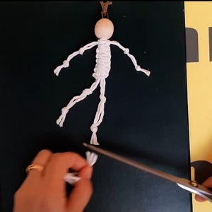 May include: A macrame doll keychain featuring a round, light-colored wooden bead head and a white knotted cord body with articulated arms and legs. The figure is attached to a metal clasp at the top, with white cord ends being trimmed by scissors.