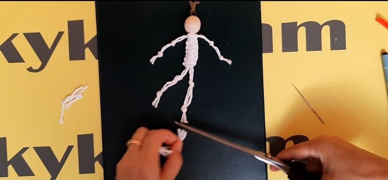 May include: A macrame doll keychain featuring a round, light-colored wooden bead head and a white knotted cord body with articulated arms and legs. The figure is attached to a metal clasp at the top, with white cord ends being trimmed by scissors.
