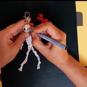 May include: A white macrame keychain shaped like a skeleton figure with a round head and knotted limbs. A grey marker is being used to color the eye socket of the skull. The keychain features a silver metal lobster clasp at the top for attachment.