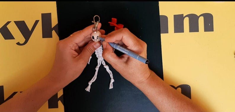 May include: A white macrame keychain shaped like a skeleton figure with a round head and knotted limbs. A grey marker is being used to color the eye socket of the skull. The keychain features a silver metal lobster clasp at the top for attachment.