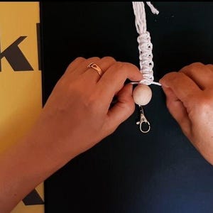 May include: A white macrame cord keychain featuring a single round light-colored wooden bead and a silver-toned metal lobster clasp. The cord is intricately knotted in a repeating pattern above the bead, with loose ends extending to the sides.