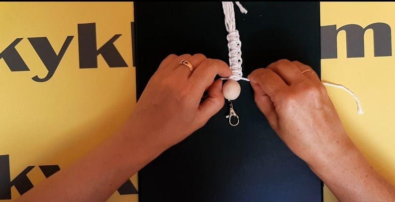 May include: A white macrame cord keychain featuring a single round light-colored wooden bead and a silver-toned metal lobster clasp. The cord is intricately knotted in a repeating pattern above the bead, with loose ends extending to the sides.