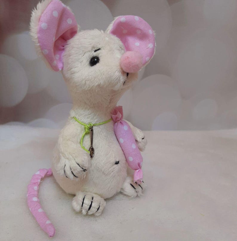 May include: A plush mouse toy with cream-colored fur, large pink ears with white polka dots, and a matching pink polka-dot tail. The mouse features a pink nose, a green string necklace with a small metal spoon charm, and holds a pink polka-dot accessory.