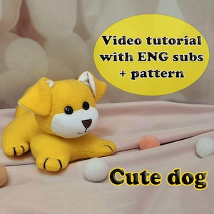 May include: A handmade yellow plush dog with black eyes and a brown nose. The dog is lying down on a pink surface. The image includes the text "Video tutorial with ENG subs + pattern" and "Cute dog".