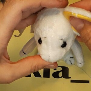 May include: A small white plush toy with black eyes and a yellow inner ear lining. The toy has a soft, textured surface and is held in place, showing its rounded head and small limbs. The design features simple stitched details on the face.