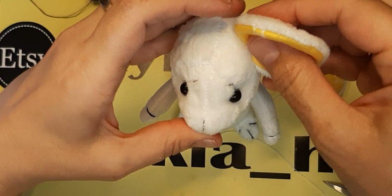 May include: A small white plush toy with black eyes and a yellow inner ear lining. The toy has a soft, textured surface and is held in place, showing its rounded head and small limbs. The design features simple stitched details on the face.