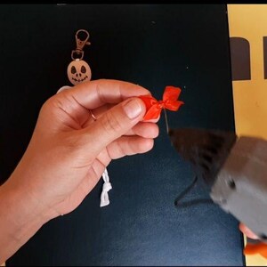 May include: A white skeleton keychain with a skull head and a metal lobster clasp. A small red ribbon bow is being attached to the skeleton using a grey hot glue gun. The skeleton has a white cord body and a circular white head with black features.