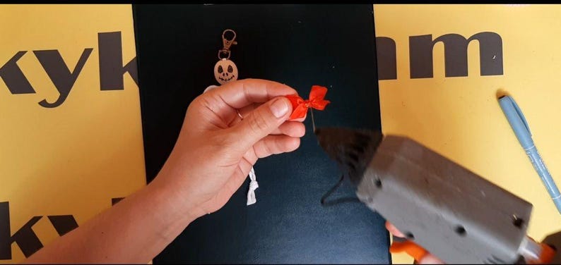 May include: A white skeleton keychain with a skull head and a metal lobster clasp. A small red ribbon bow is being attached to the skeleton using a grey hot glue gun. The skeleton has a white cord body and a circular white head with black features.