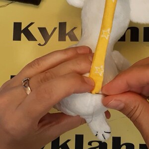 May include: A white plush toy being hand-stitched with a needle and thread. A yellow fabric strip featuring a white star pattern is being attached to the back of the plush toy. The toy has visible white ears and a small white tail with black stitching.
